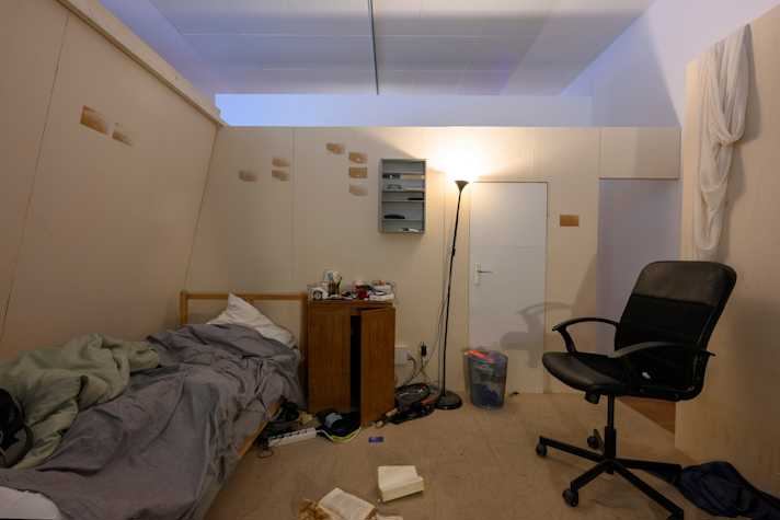 A bedroom space with white walls filled with a bed, a computer chair and storage furniture. On the floors sits open books and cables. 