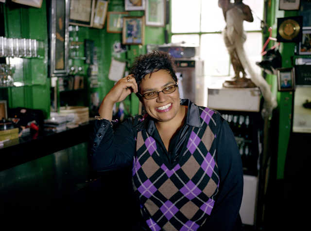 Woman brightly smiling in bar wearing diamond patterned sweater. There are many photography on the walls in the background which is out of focus