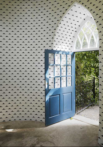 An open blue door in a small arched alcove opens onto a curved room. The white walls are painted with a repeating pattern of curved black triangles. Outside this door a garden is just visible along with a black cast iron railing. Sunlight streams through the door and from a window above it.