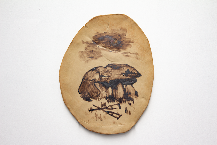 An oval ceramic plate on a white wall. The ceramic is a tan brown colour with an inky black drawing of a monk bending over a stone dolman in prayer. In front there are two crutches in the grass, crossed over each other. Above, some clouds and a round moon are visible.