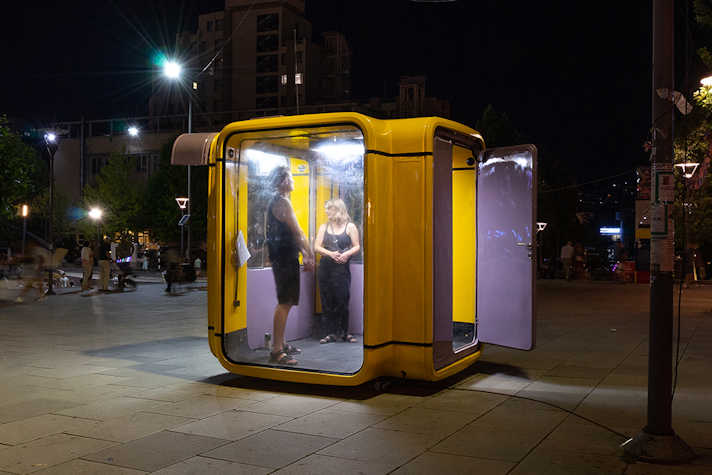 A photo of an outdoor urban space at night. On the pavement is a large cube structure made with glass walls, a yellow metal frame, and a purple door which is open. Two people stand inside it. 