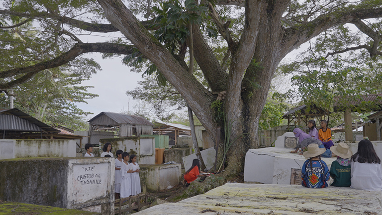 A photo of a graveyard with a large tree at the centre and a collection of small wooden buildings in the background. A group people stand beside one of the graves. 