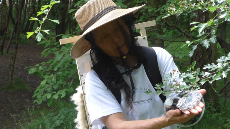 A woman waring a sunhat and insect net sits in outside in an area surrounded by trees. In her hands she holds a machine which is connected to a tube.
