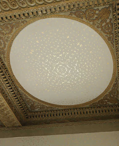 A photograph of a ceiling replete with an ornate circular alcove bordered with golden floral-styled cornicing. In the centre there is a symmetrical design in gold leaf. The shapes that make up the pattern resemble abstract flowers or avian forms.