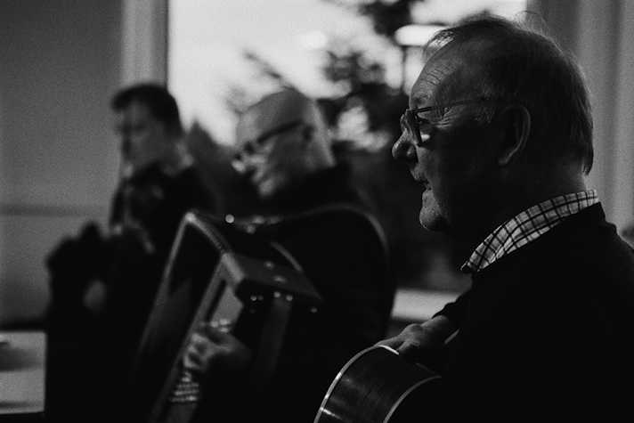 B+W image of an older white man holding a guitar in profile and in sharp focus, another white male behind him playing the accordion in medium focus, a third white male behind him playing the violin out of focus.]