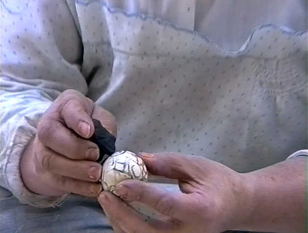 Video still, a close up of a woman’s hands drawing circles an egg in black charcoal. The woman wears a white dress with blue spots.