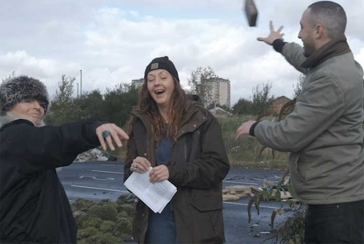 Three people stand outside in a carpark area filled with rubble and foliage. In the background there is a high rise flat.
