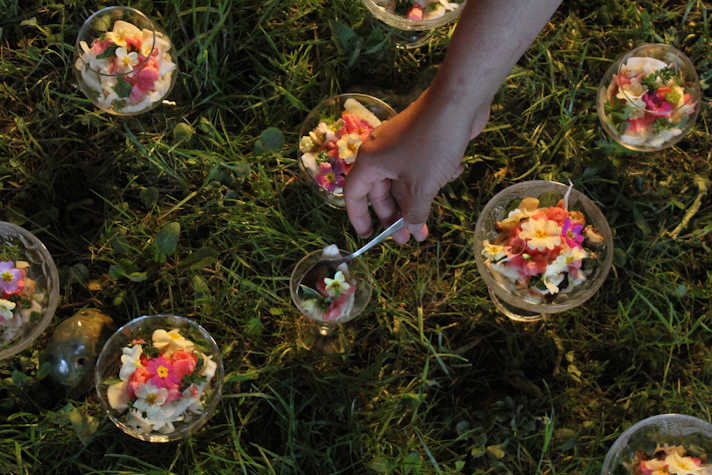 A photo taken from above showing an area of grass covered by drinking glasses containing flowers. A hand holding a spoon reaches into the centre glass. 