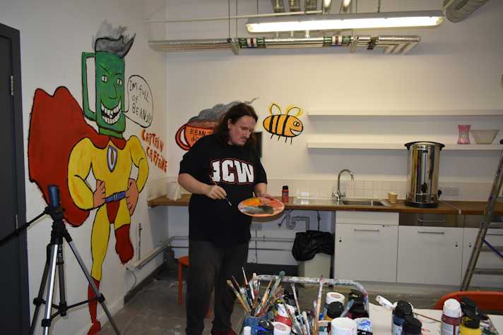 A photograph of a man standing in a kitchen area painting a mural on a white wall. The painting depicts a superhero figure wearing a yellow suit and red cape with a green mug as a head. Text next to the painting reads 'captain caffeine'.