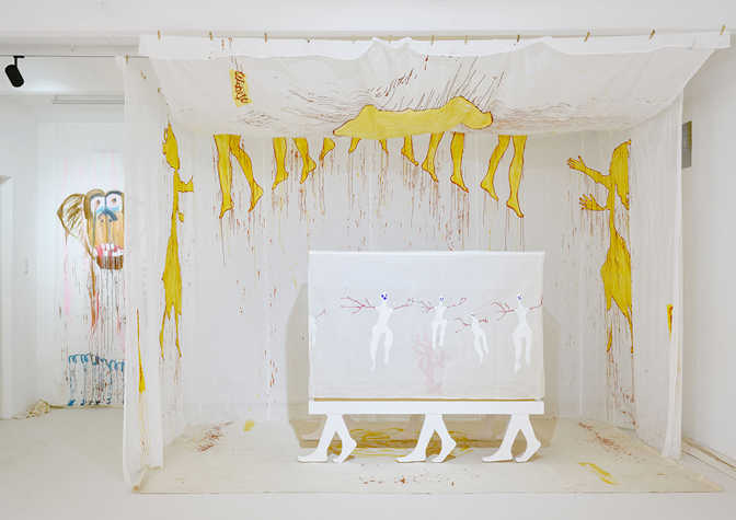 A white room with white fabric hanging from the ceiling. On the walls outlines of human figures are painted. In the centre of the room is a canvas on a frame held up by 6 legs resembling human legs.