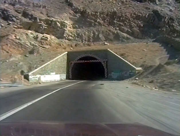 Video still taken from the front of a red car driving towards a tunnel opening. Sandy coloured mountains loom overhead.