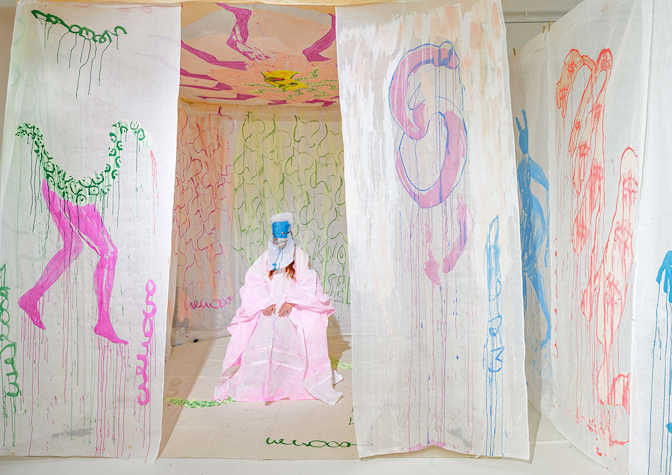 A room with white walls and white fabric hanging from the ceiling which are painted with brightly coloured shapes and body parts. At the centre of the room sits a person in a long pink dress with red hair. Their face is covered by a blue and white mask.  