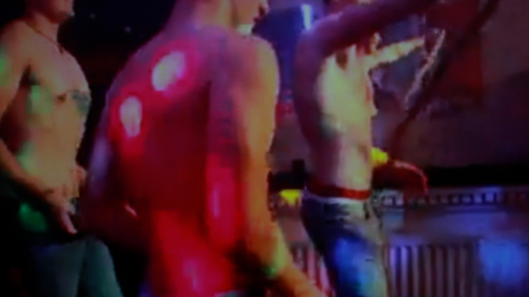 Three topless bodies with traditionally 'male' physiques with toned muscles are dancing in a club, the image is quite pixelated suggesting it is archival