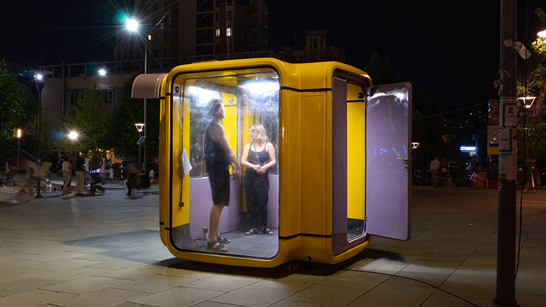 A photo of an outdoor urban space at night. On the pavement is a large cube structure made with glass walls, a yellow metal frame, and a purple door which is open. Two people stand inside it. 