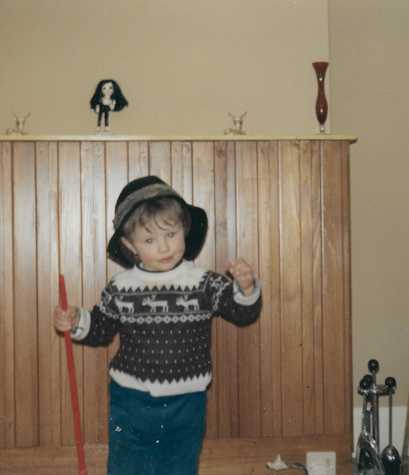 A photo of artist Delaine Le Bas as a two year old child. Pictured standing in front of a wooden backdrop, with a red stick in their right hand.