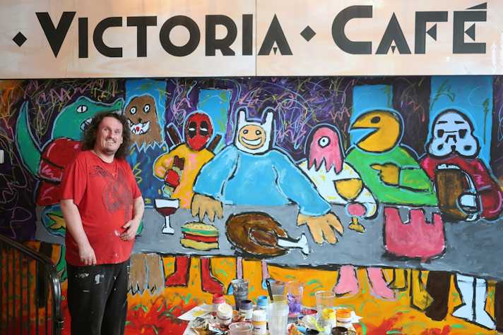 A photo of a man standing in front of a mural painted on a wall which depicts five characters standing behind a grey table. The characters from left to right, Chewbacca (Star Wars), Wade Wilson (Deadpool), Finn the Human (Adventure Time), Zoidberg (Futurama) and Pac Man. On the table there is a wine, a burger, and a roast chicken. The back ground is blue and purples with squiggle lines in the purple sections. Across the top of the image is a banner with the text 'Victoria Cafe'.