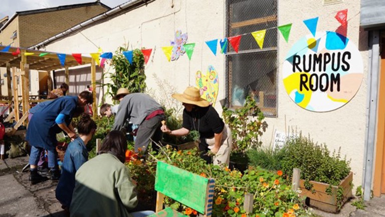 A photograph featuring a group of people gardening in planters outside of a building. The building has a circular logo sign with the text 'Rumpus Room'.