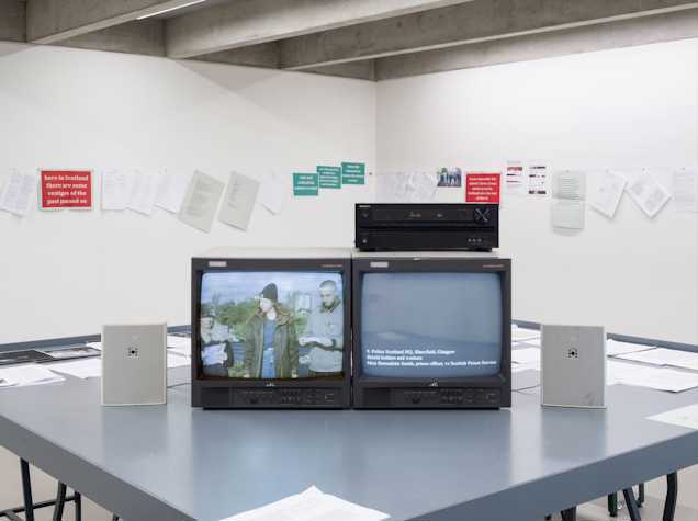 A photo of a room with a grey table with two TVs sitting on it, the left screen has three people standing outside and the right screen is black with white text reading 'Police Scotland HQ, Shawfield, Glasgow.'  In the background of the image are white walls covered by a series of signs and pieces of paper. 